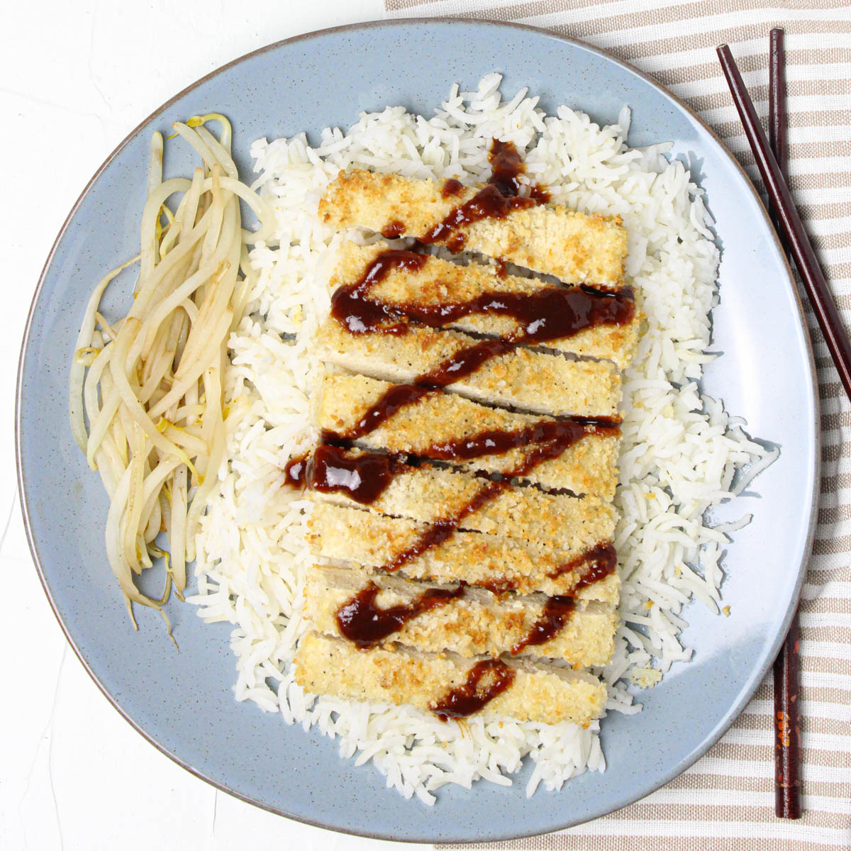 Blue plate with rice, bean sprouts, and tofu katsu with drizzled sauce.
